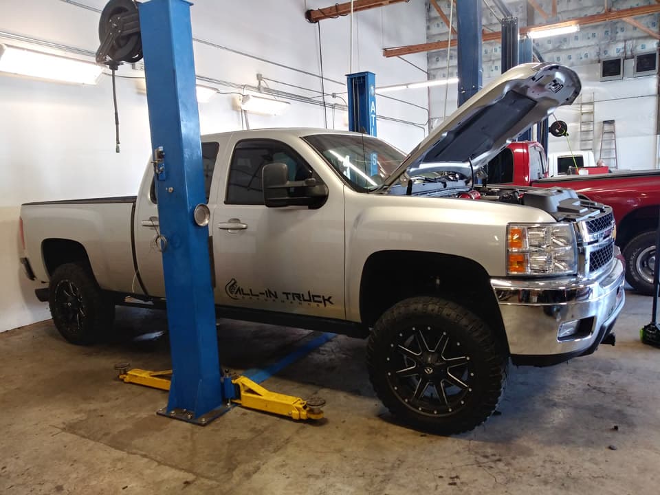 A silver Chevrolet Silverado up on a two-post lift with yellow lift arms, hood open, in the lit shop bay.