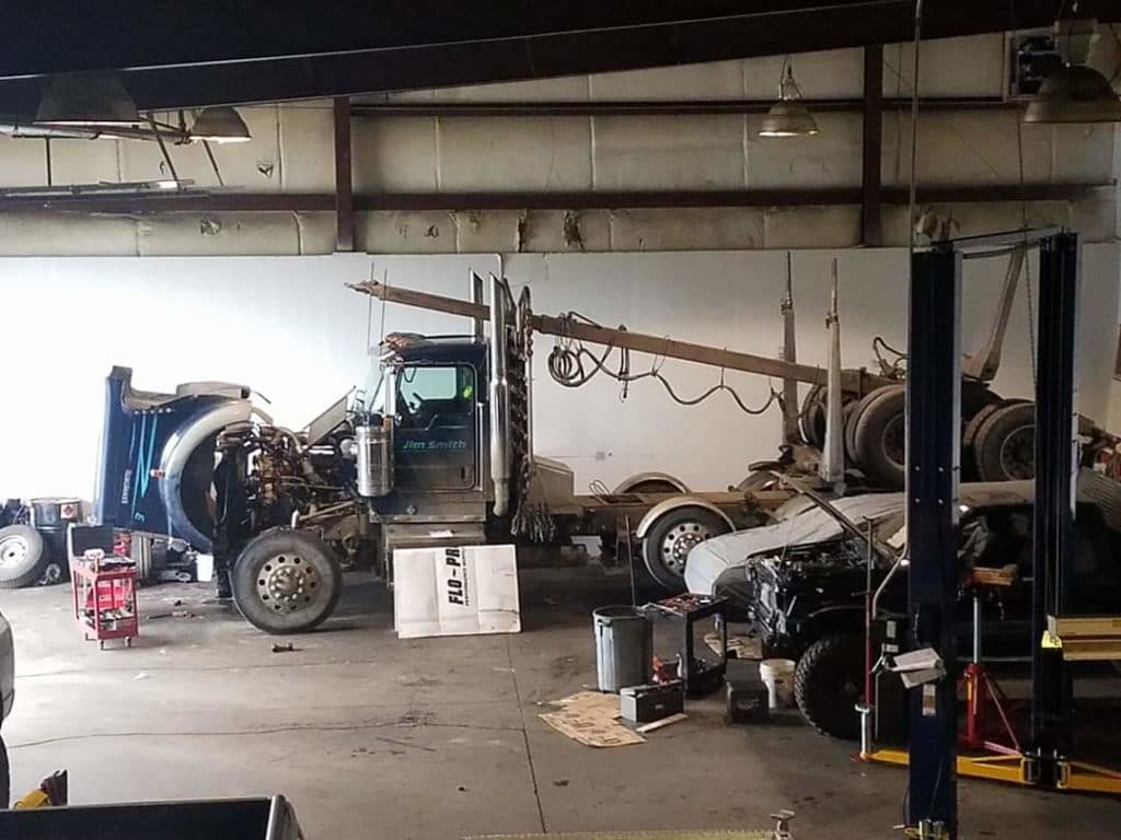 A semi tractor with its cab tilted forward for engine access, next to a pickup on a lift.